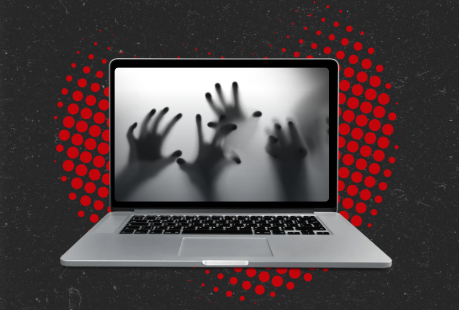 A gray laptop on a black background with red dots. Hands reach out of the laptop screen in a spooky way.